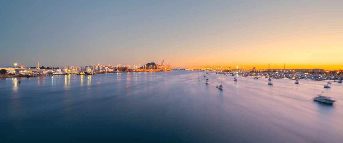 Panoramic view of Tauranga Harbour at sunset with Mount Maunganui, port cranes, and marina lights — The Gold House® precious metals brokerage, New Zealand