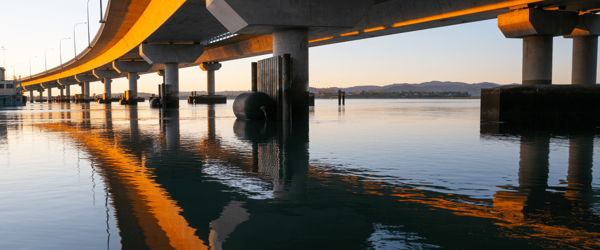 Sunrise over a Tauranga harbour bridge with calm water reflections — The Gold House® Trading Desk for gold, silver, and platinum bullion, New Zealand