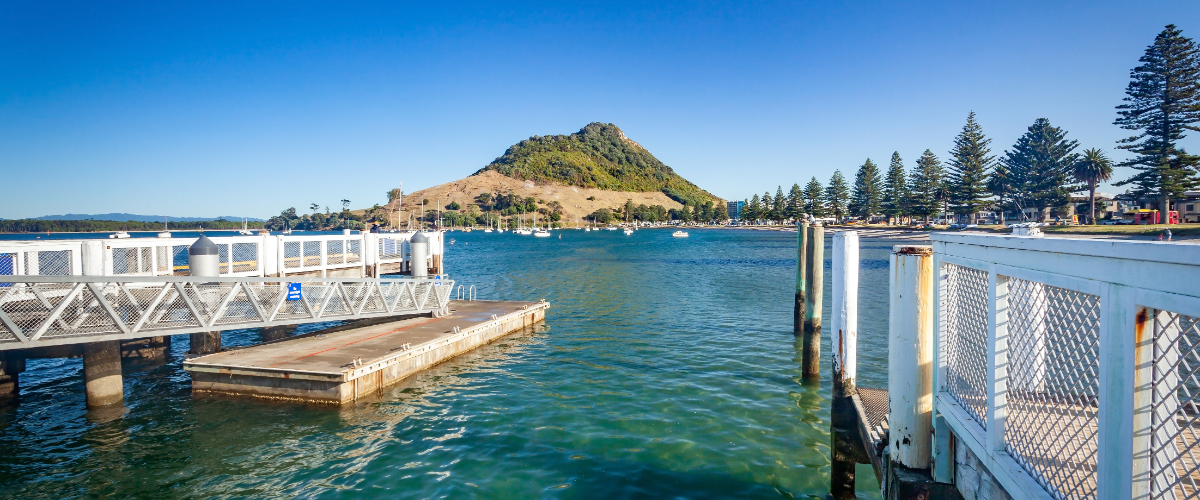 Bright marina view of Mount Maunganui (Mauao) across turquoise harbour water with pier and sailboats — The Gold House® mint and refinery bullion, Bay of Plenty, New Zealand