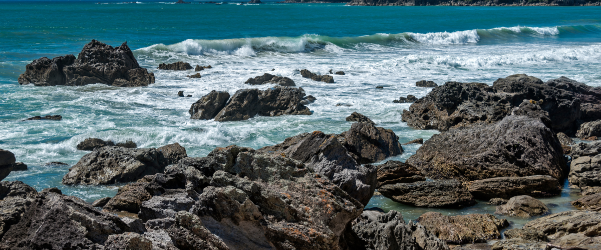 Turquoise surf breaking over dark volcanic rocks on the Bay of Plenty coast — The Gold House® platinum bullion products, New Zealand