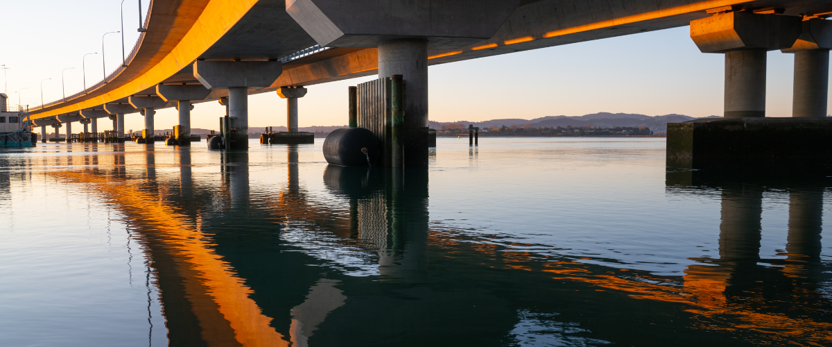 Sunrise over a Tauranga harbour bridge with calm water reflections — The Gold House® Trading Desk for gold, silver, and platinum bullion, New Zealand