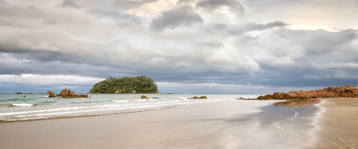 Wide sandy beach with Moturiki Island offshore under a soft overcast Bay of Plenty sky — The Gold House®, New Zealand